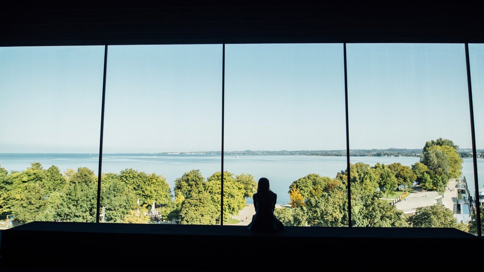 Das Panoramafenster im vorarlberg museum mit Blick auf den sommerlichen Bodensee und die Silhouette einer sitzenden Frau