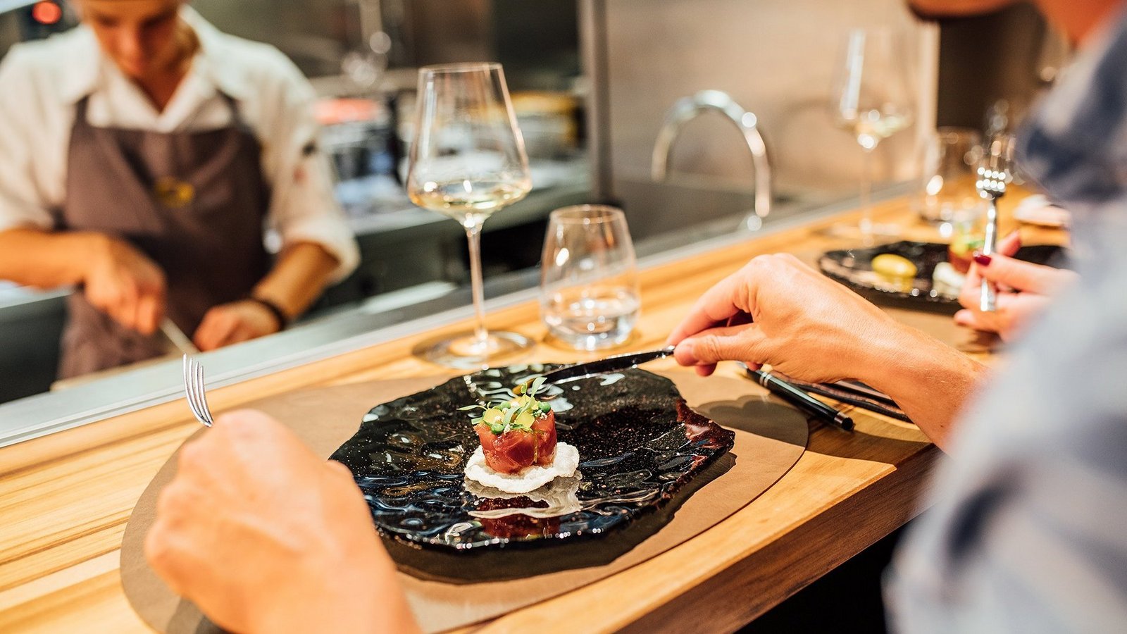 Eine Person sitzt vor einem angerichteten Essen mit direktem Blick in die Küche des Restaurants
