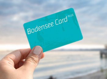 Eine Frau hält die türkise Bodensee Card PLUS, im Hintergrund der See