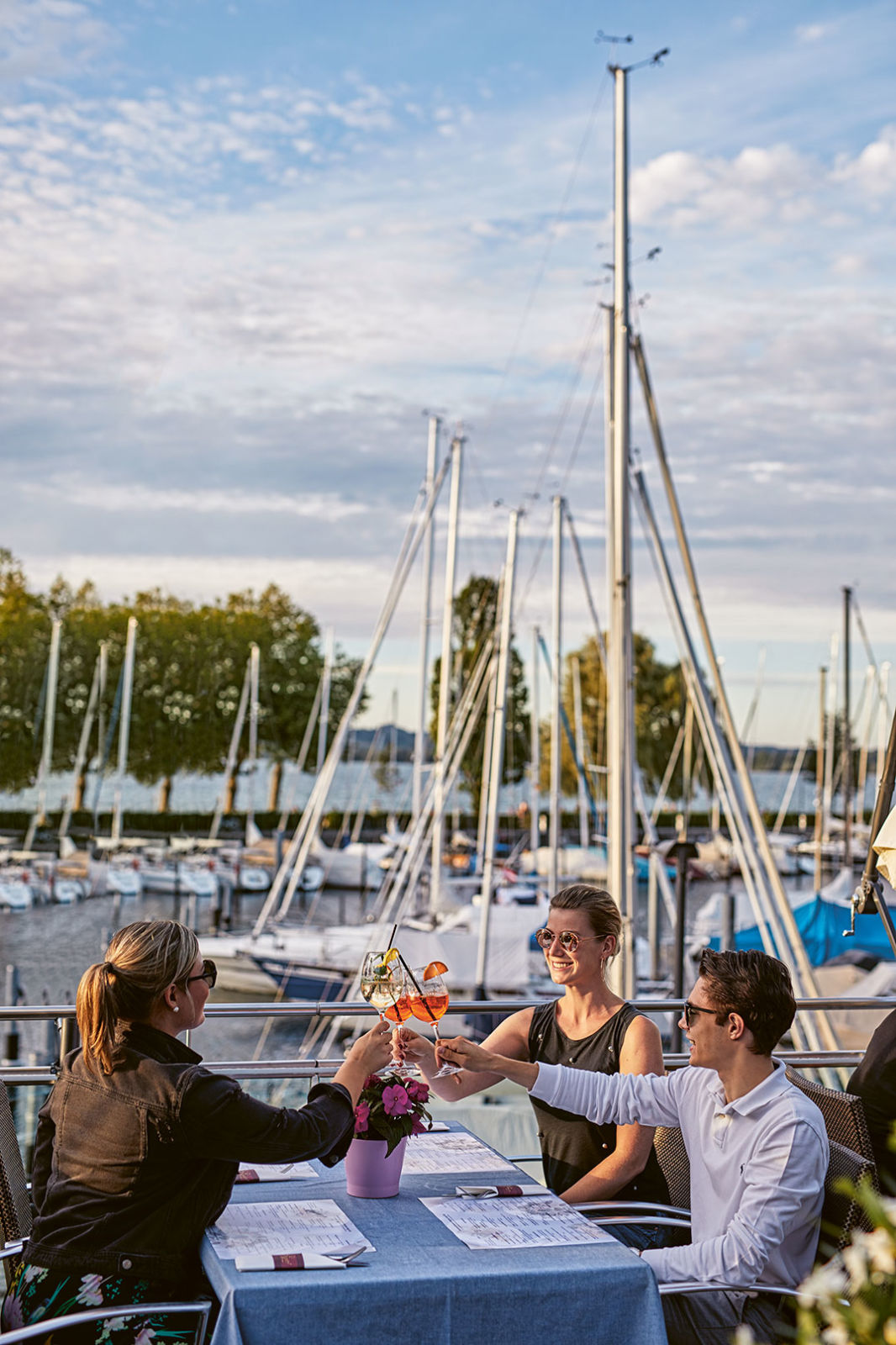 Drei Junge Leute stoßen mit ihren Drinks an. Im Hintergrund ein Hafen mit Segelbooten.